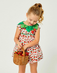 Toddler girl in a strawberry-print romper with a green leaf collar, holding a wicker basket filled with fresh strawberries.