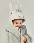 Young child wearing a pale pink wool unicorn hat with a silver mane and satin ribbon ties, bundled up in a cozy grey coat with faux fur trim