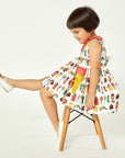 Girl in cotton Two Scoops Dress with The Very Hungry Caterpillar print, coral collar, and quilted ice cream pockets, sitting on chair with playful pose