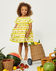 A young girl in a striped yellow dress with lemon patterns stands barefoot at a fruit market, holding a mini cart of lemons