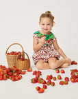 Smiling toddler wearing a strawberry-print romper, sitting with fresh strawberries and a wicker basket on a white background