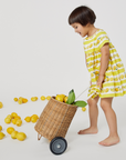 Young girl in a lemon-print dress playfully pulling a wicker cart filled with lemons, surrounded by scattered lemons.