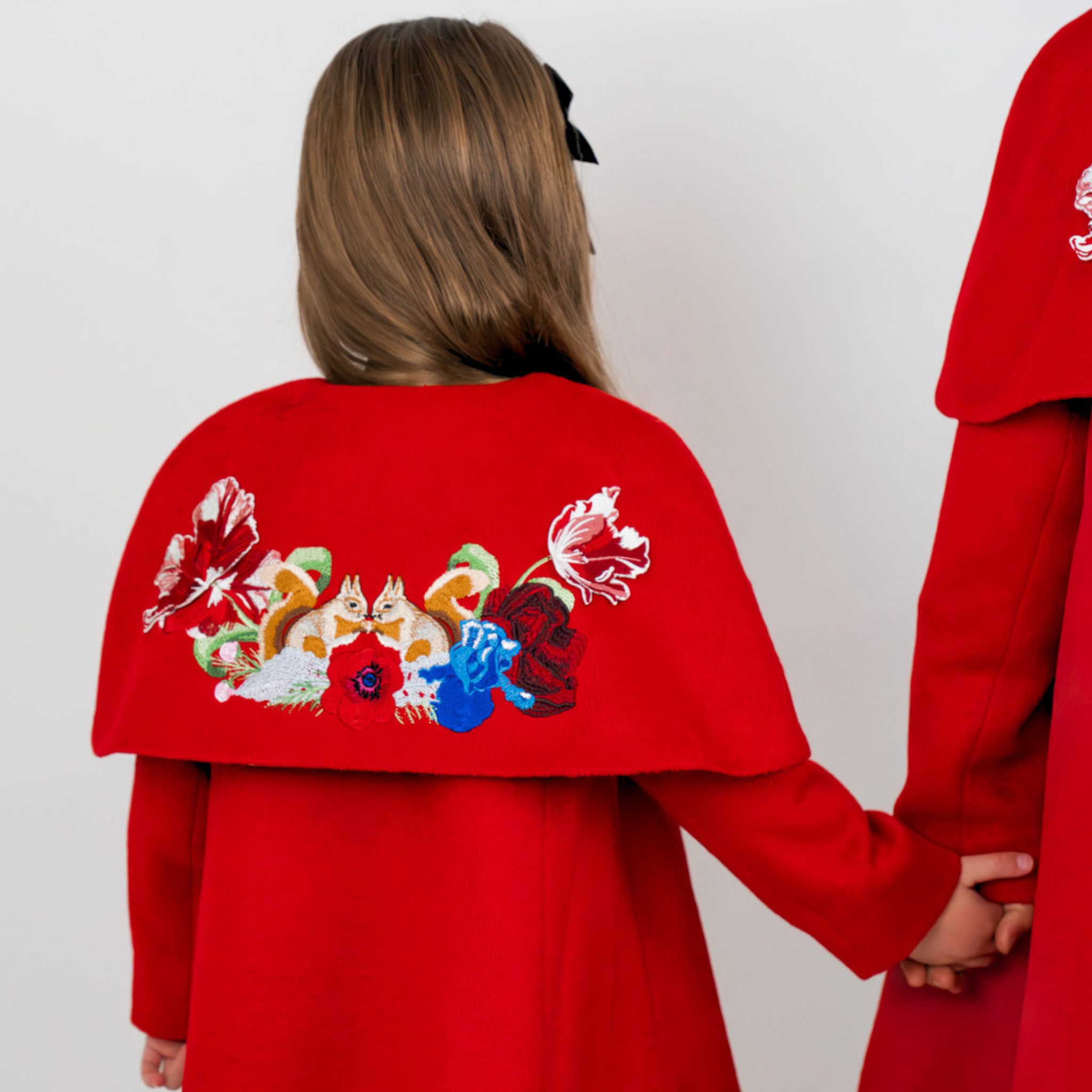 A child wearing a bright red cape-style coat, shown from the back, holding hands with another person in a matching red coat. The back of the coat features a large embroidered floral design wiith two squirrels  in white, blue, green, and red.