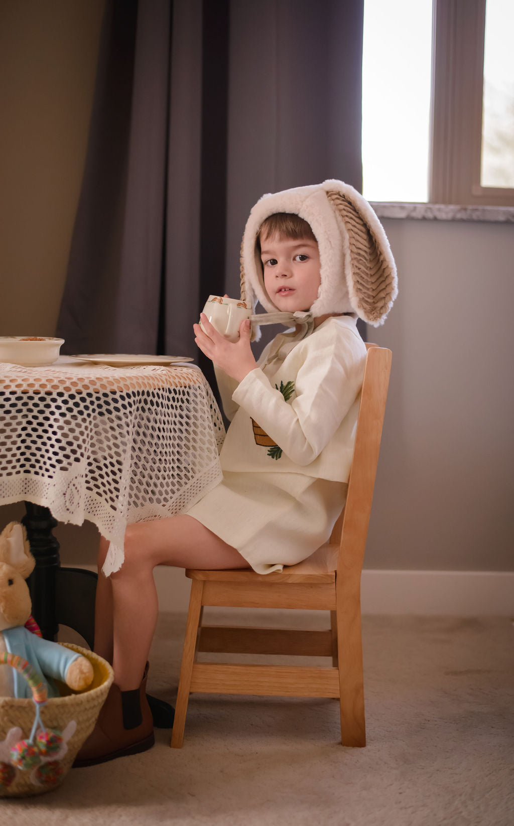 Child wearing Little Goodall embroidered bunny linen jacket and dress, sipping tea while wearing a plush bunny bonnet