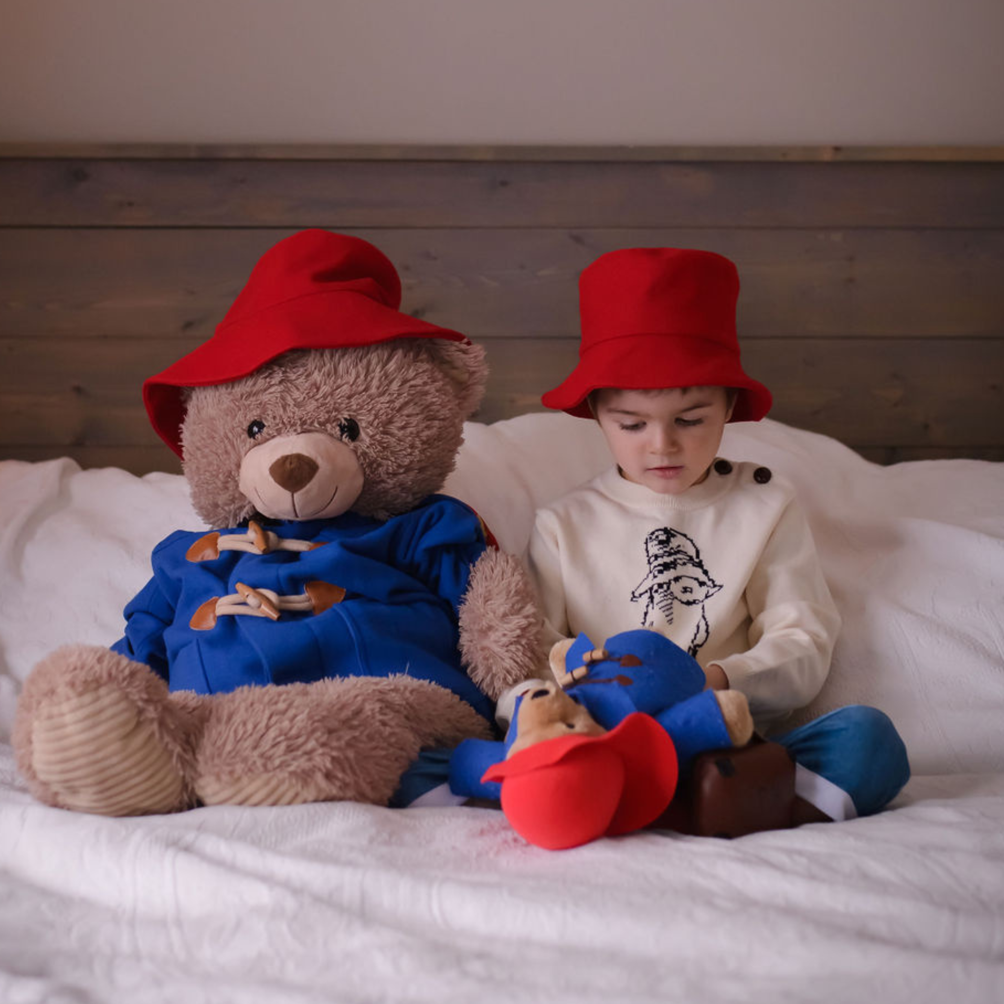Child and Paddington soft toy sit on a bed wearing matching red bucket hats, with the child playing with plush toys.