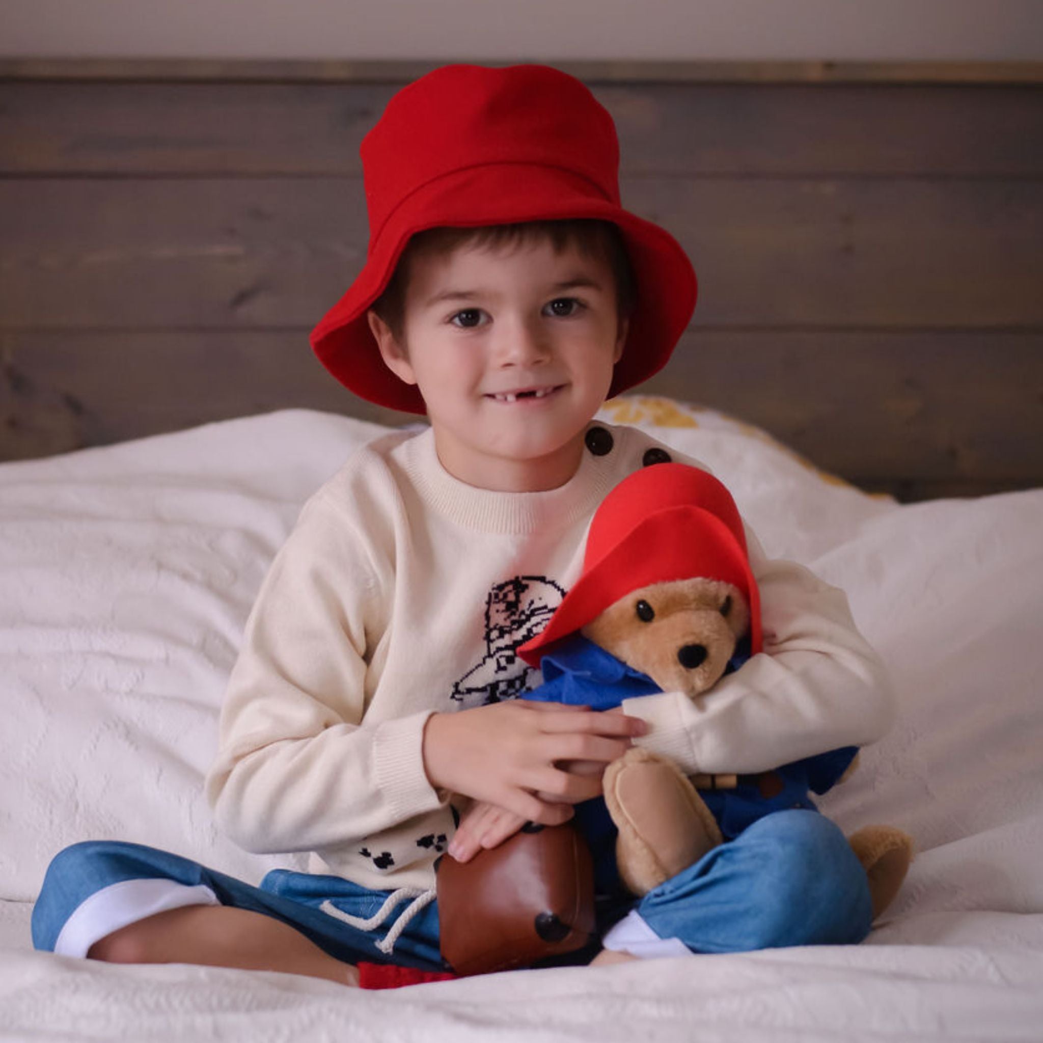 child wearing a red bucket hat and a Paddington Bear sweater sits on a bed, hugging a Paddington Bear plush toy dressed in blue and red.