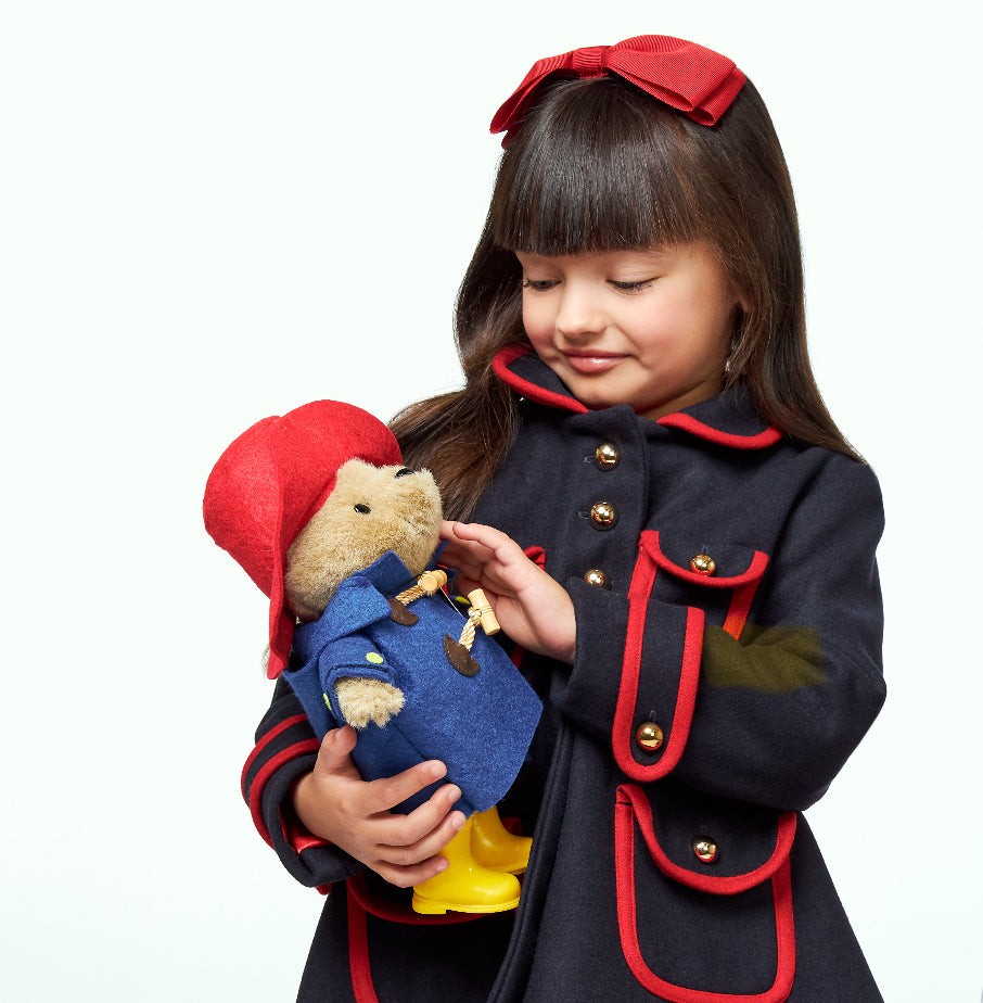 A girl in a dark blue coat holds the Classic Paddington Bear 10" Soft Toy, dressed in a blue toggle coat, red hat, and yellow boots.