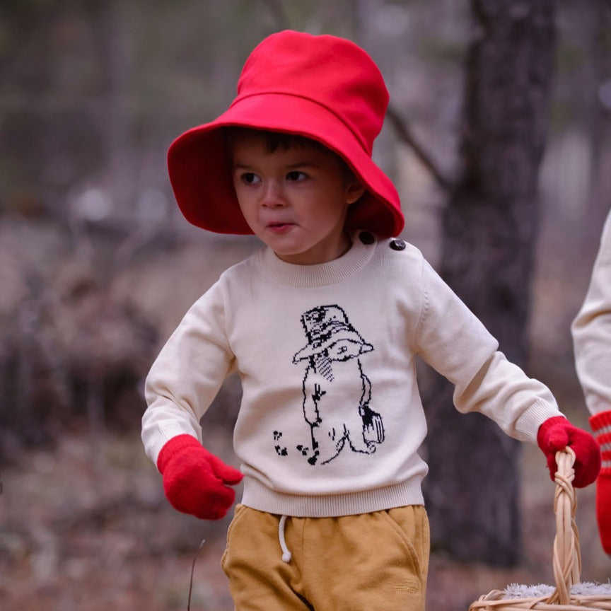 child wears an ivory knit sweater with a black embroidered Paddington Bear design, ribbed cuffs, and button details, pants,a red hat, and gloves,
