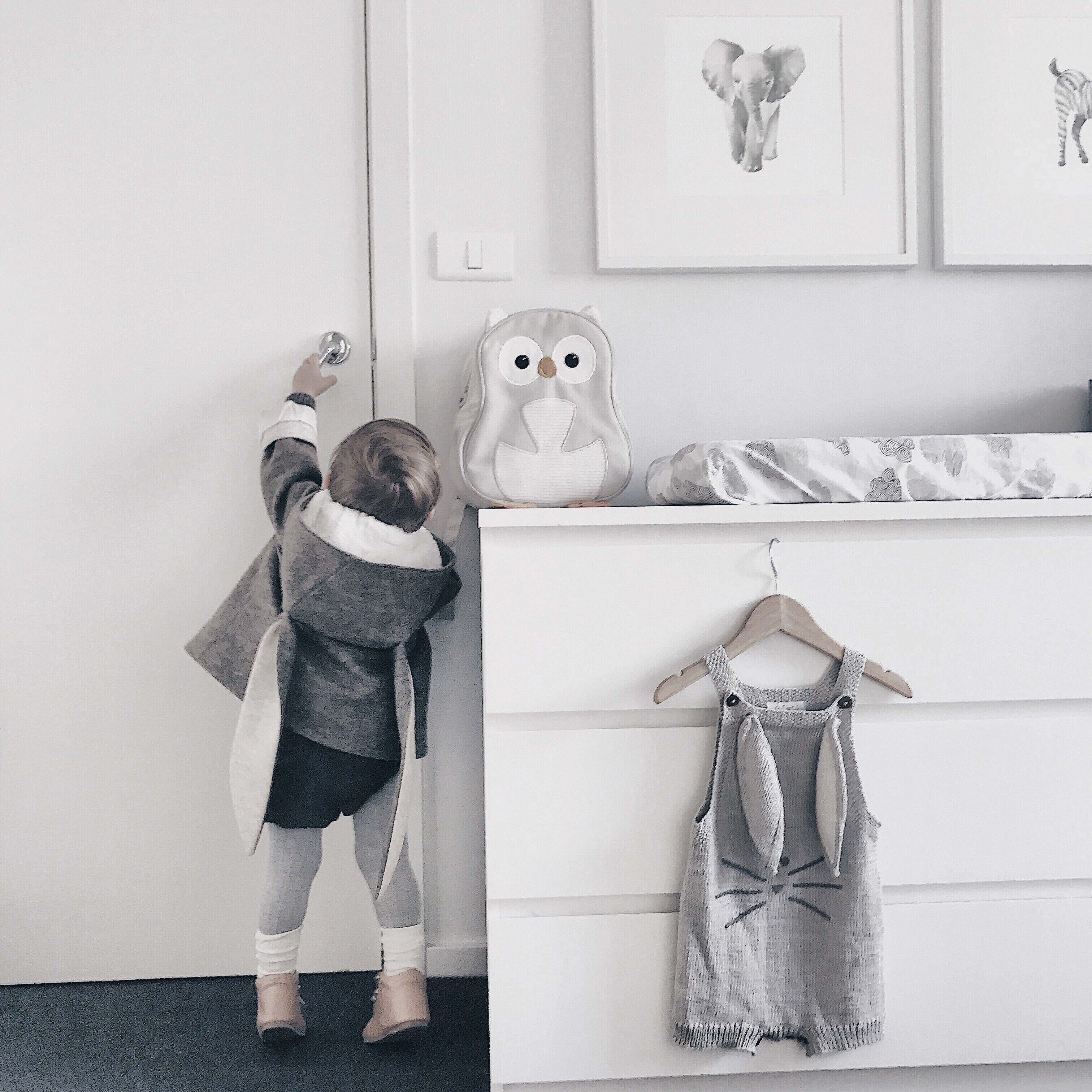Toddler wearing the Flopsy Bunny Coat with floppy-eared hood, reaching for a door handle in a softly lit nursery.