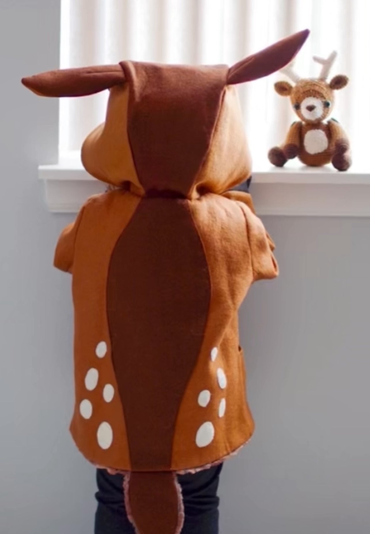 Toddler wears the Frolicsome Fawn Coat, showing its spotted design, long deer tail, and hood with ears. A plush fawn toy sits on the windowsill