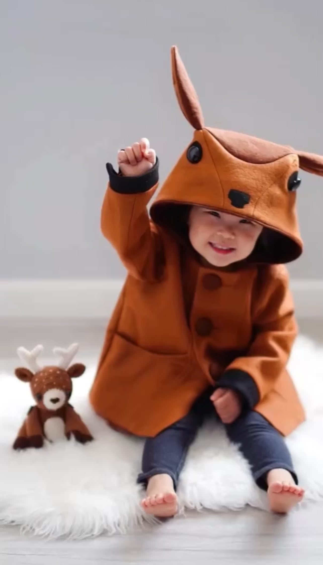 Toddler wearing the Little Goodall Frolicsome Fawn Coat, smiling and raising a hand, with deer hood ears perked up and a plush fawn toy beside them