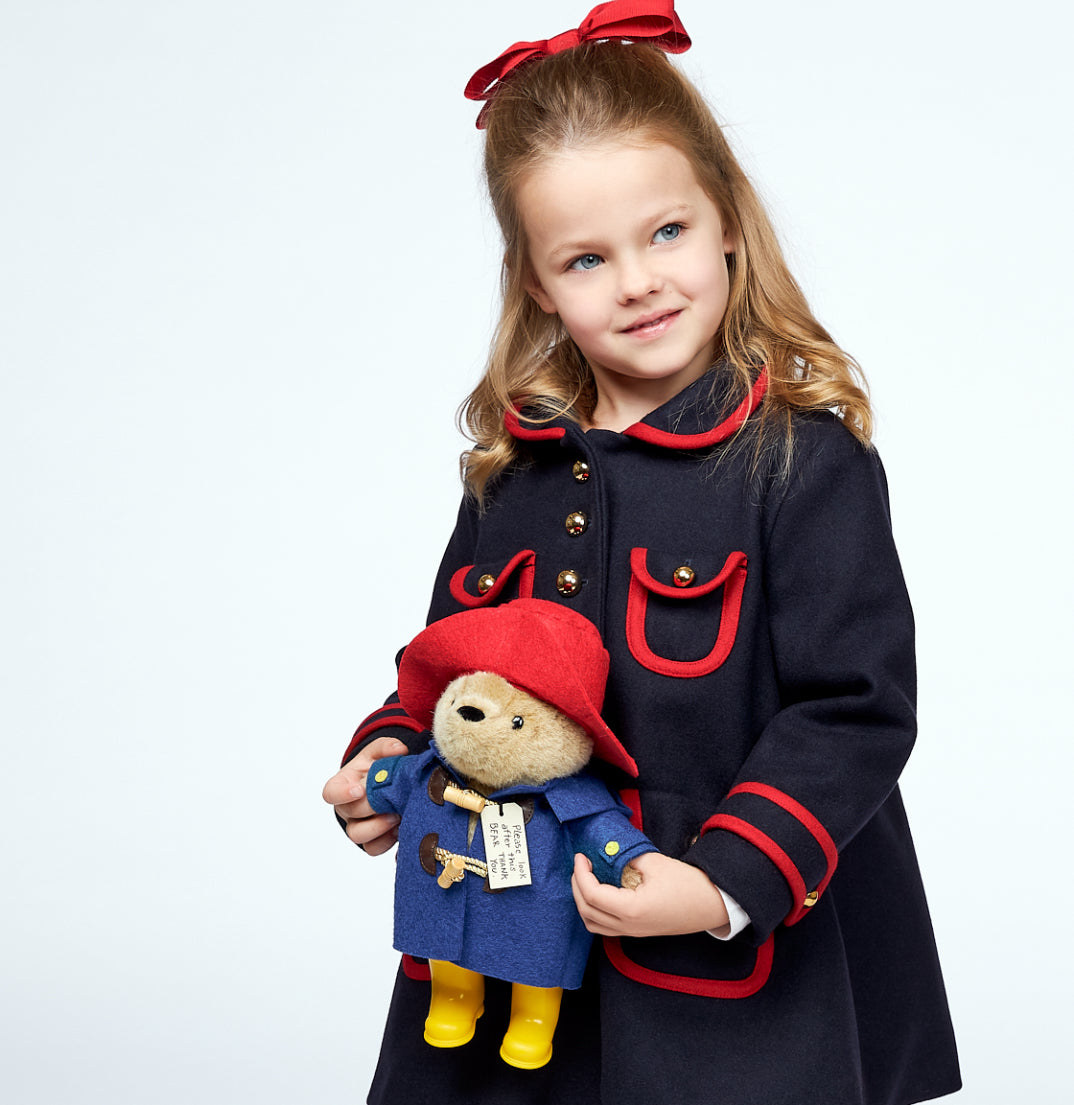Young girl in a dark blue wool coat with gold buttons holds a Paddington Bear soft toy, featuring a red hat, blue wool coat, and yellow boots