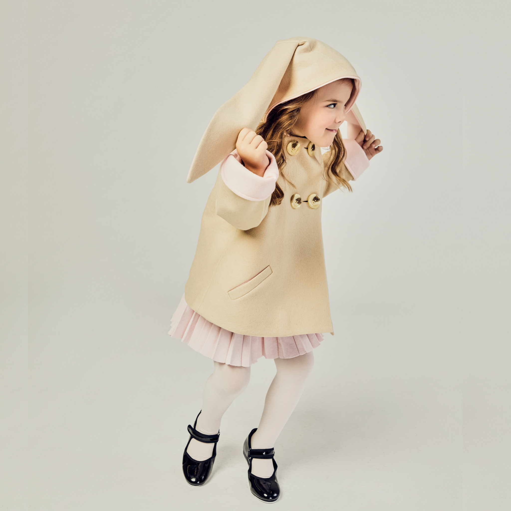 Young girl playing in Luxe Bunny Coat in Sand & Pink, featuring long bunny ears, wool fabric, and pink-lined cuffs.
