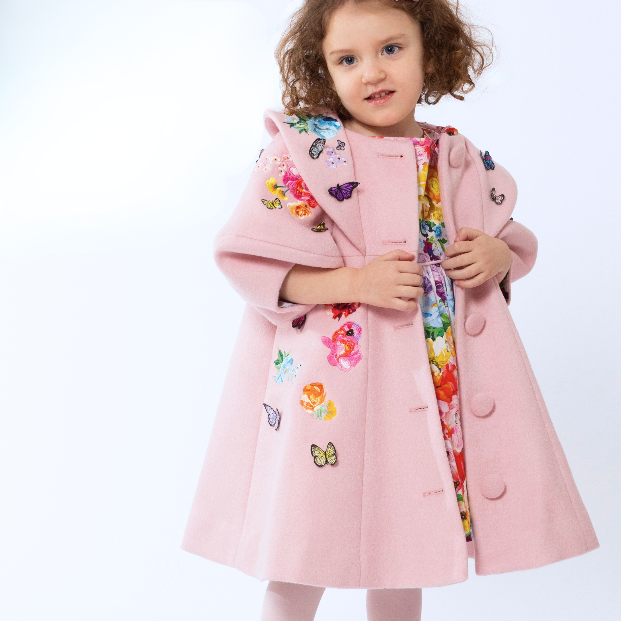 Young girl wearing a pink wool coat with floral embroidery, butterfly appliqués, and a vibrant floral dress underneath