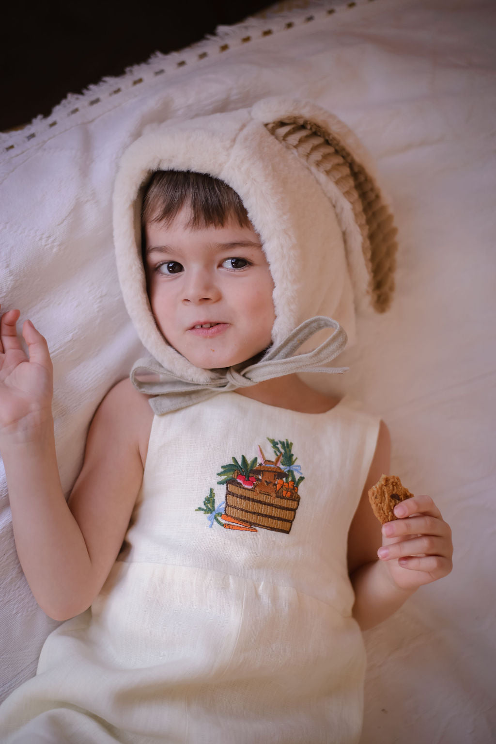 Ivory linen romper with embroidered bunny in a basket, adjustable straps, and button closures. Gender-neutral baby and toddler outfit