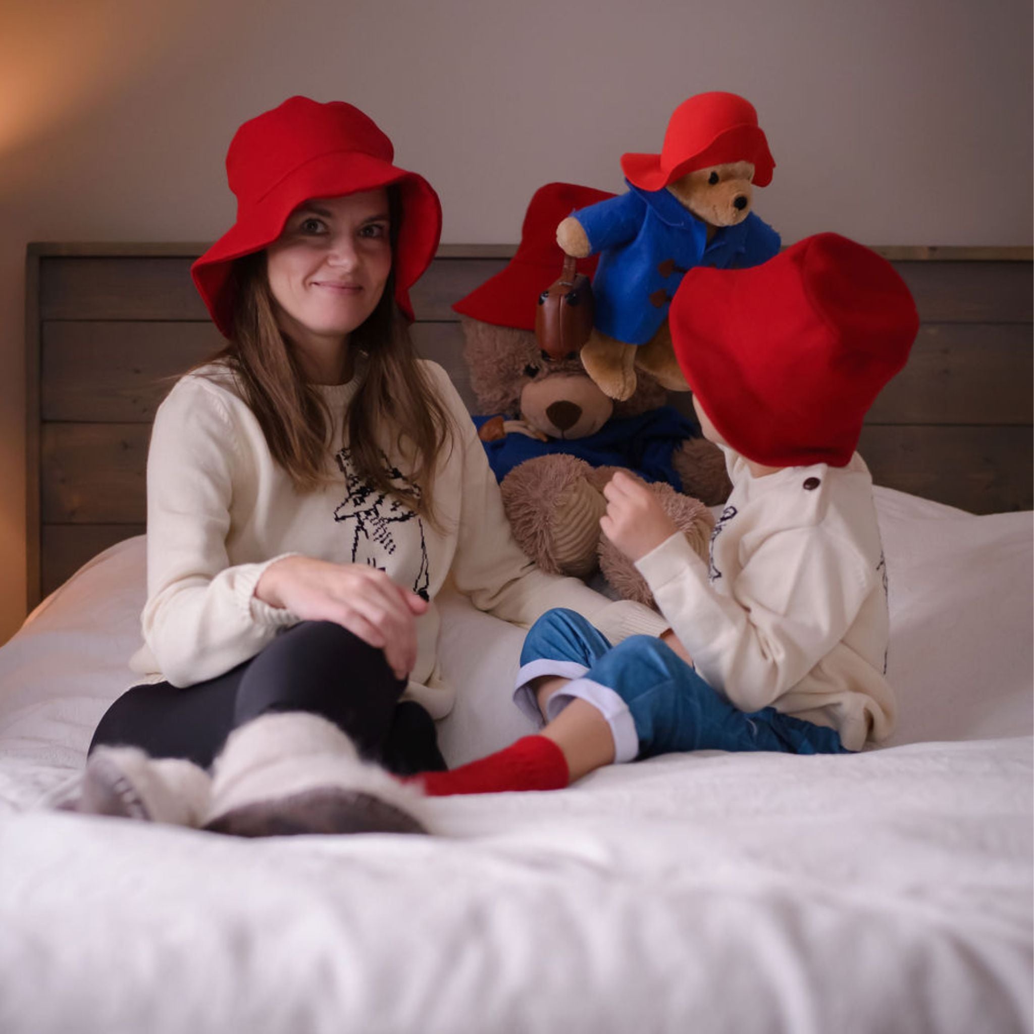 Mother and child wearing matching red wool Paddington bucket hats, sitting on a bed with plush Paddington Bear toys