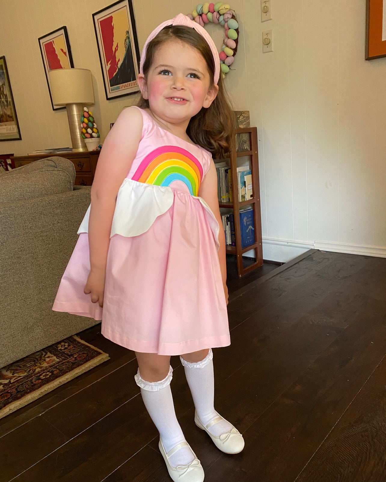 Young girl wears a pink sundress with a rainbow bodice and cloud peplum, paired with knee-high socks and white shoes, posing indoors with pastel decor