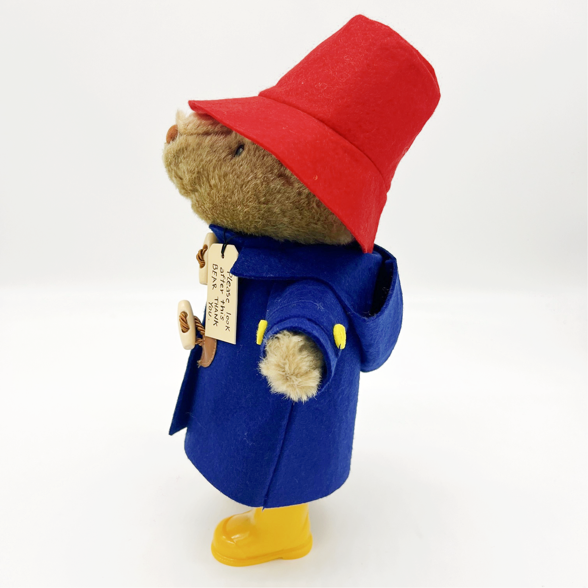 Side view of Paddington Bear 10" plush toy in a blue toggle coat, red hat, and yellow boots with "Please Look After This Bear" tag