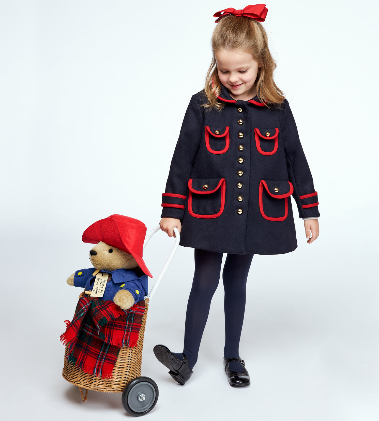 girl wearing a navy wool coat with red trim, gold buttons, and a matching red bow pulls a wicker cart carrying a 16-inch Paddington Bear plush toy
