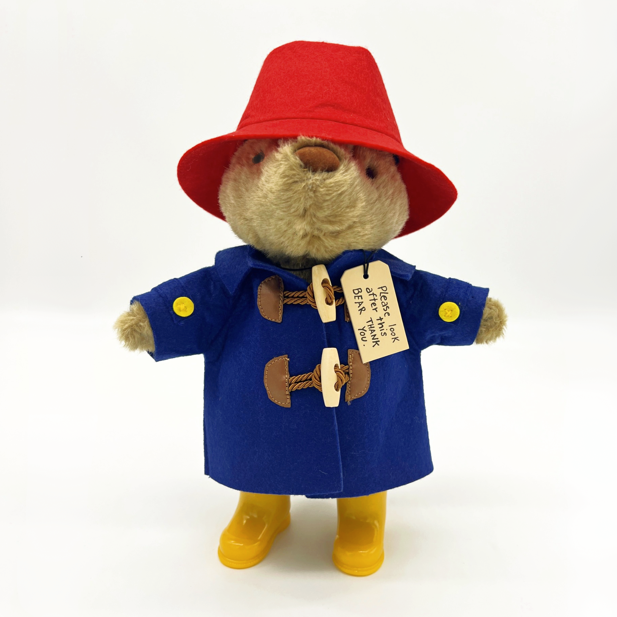 Paddington Bear plush in a blue duffle coat, red hat, yellow boots, and his iconic “Please look after this bear. Thank you.” tag