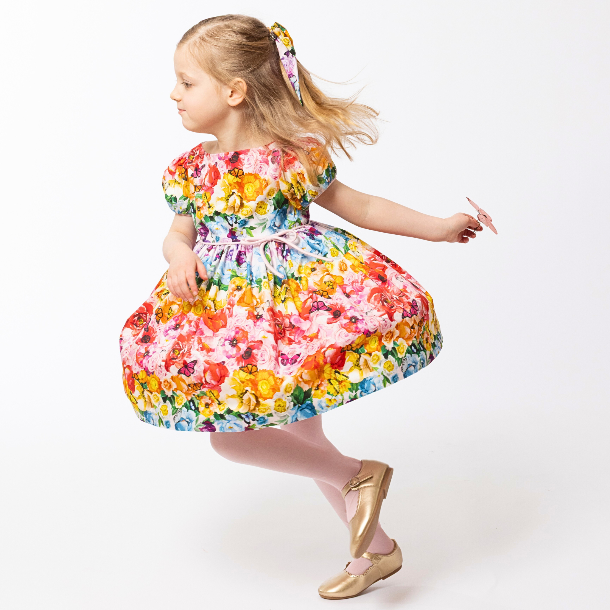Young girl twirling in the Rainbow Bloom Dress, featuring a vibrant floral and butterfly print, puff sleeves, and a tie waist. 