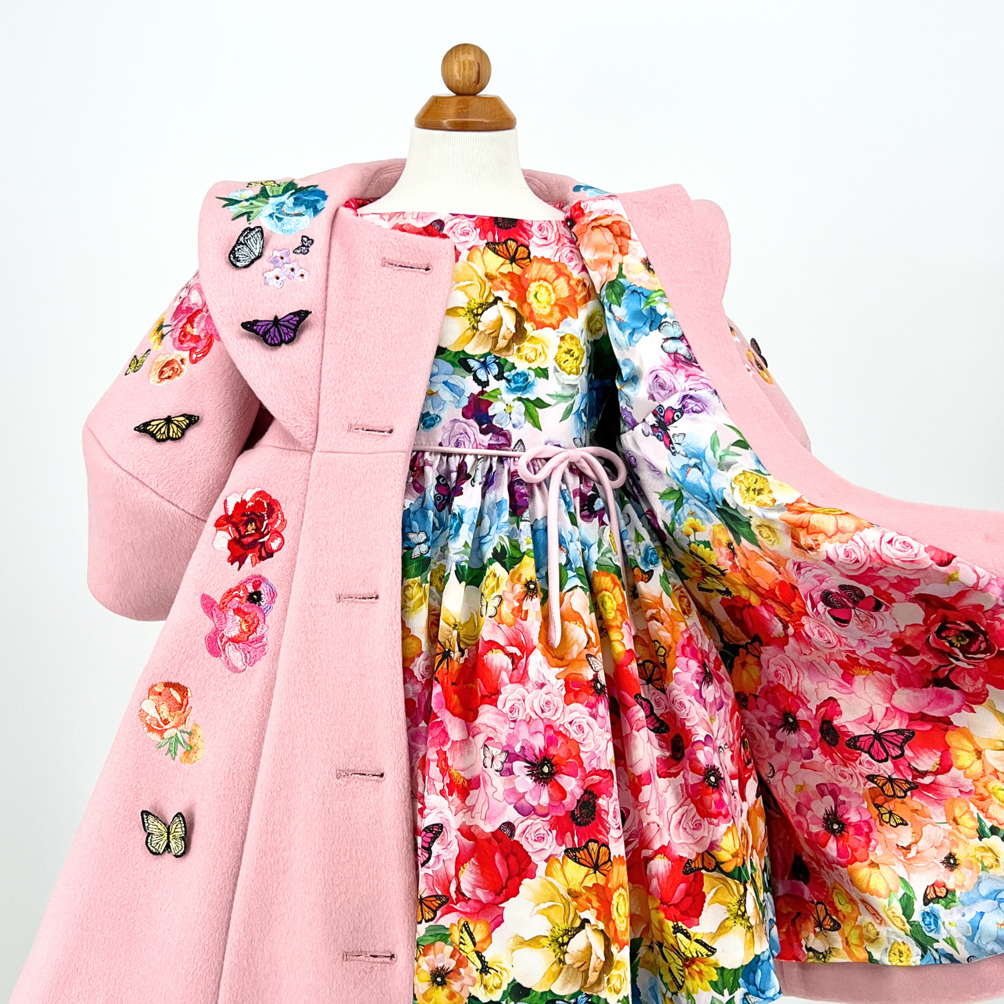 Pink wool girls' coat with embroidered flowers, butterfly appliqués, and vibrant floral lining, shown over a matching dress