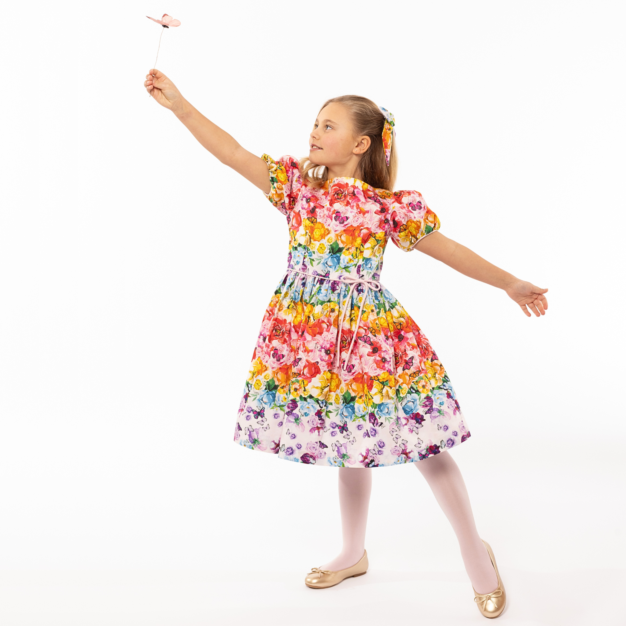 Girl in Rainbow Bloom Party Dress, holding a butterfly wand. Vibrant floral print with puff sleeves and a tie waist, perfect for special occasions