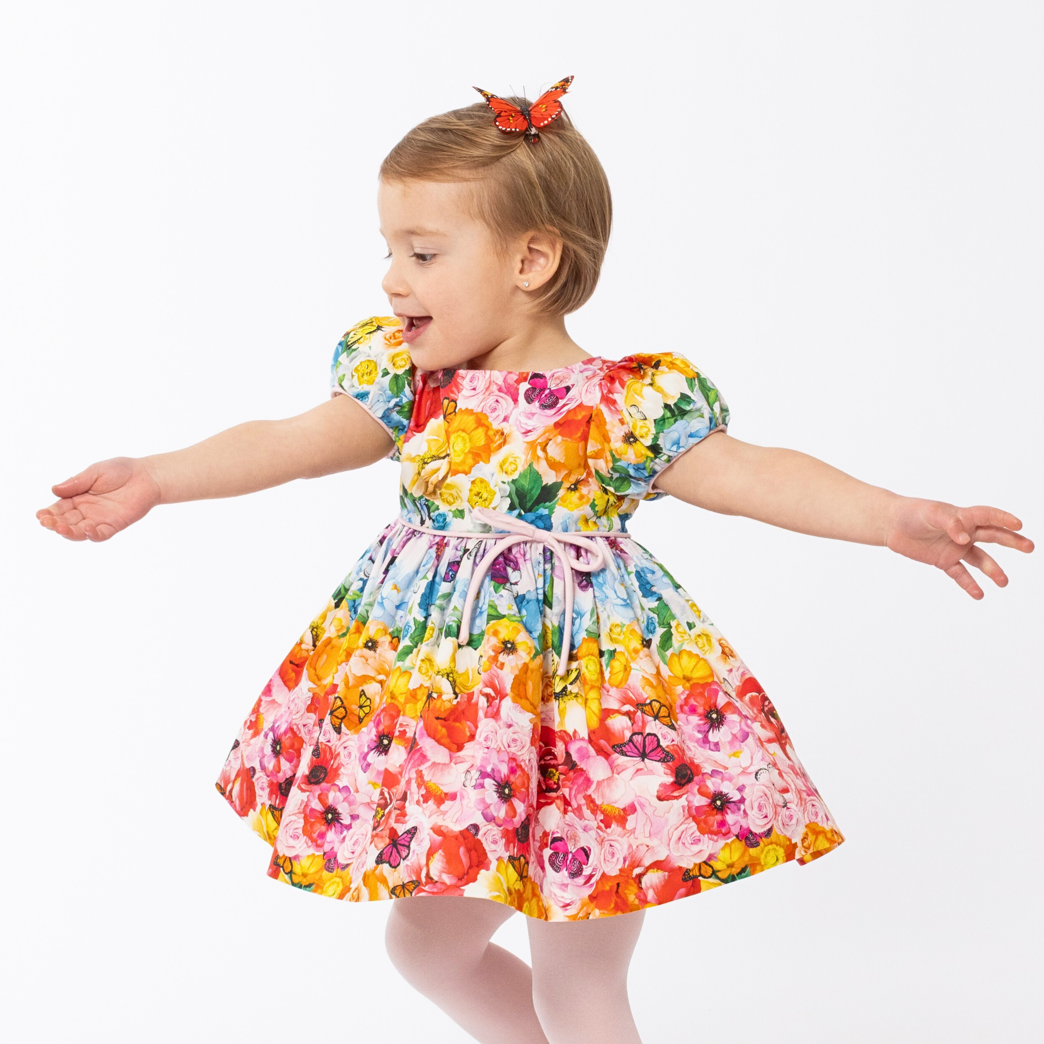 Girl twirling in a vibrant Rainbow Bloom Party Dress with puff sleeves, a pink waist bow, and a floral butterfly print, perfect for special occasions
