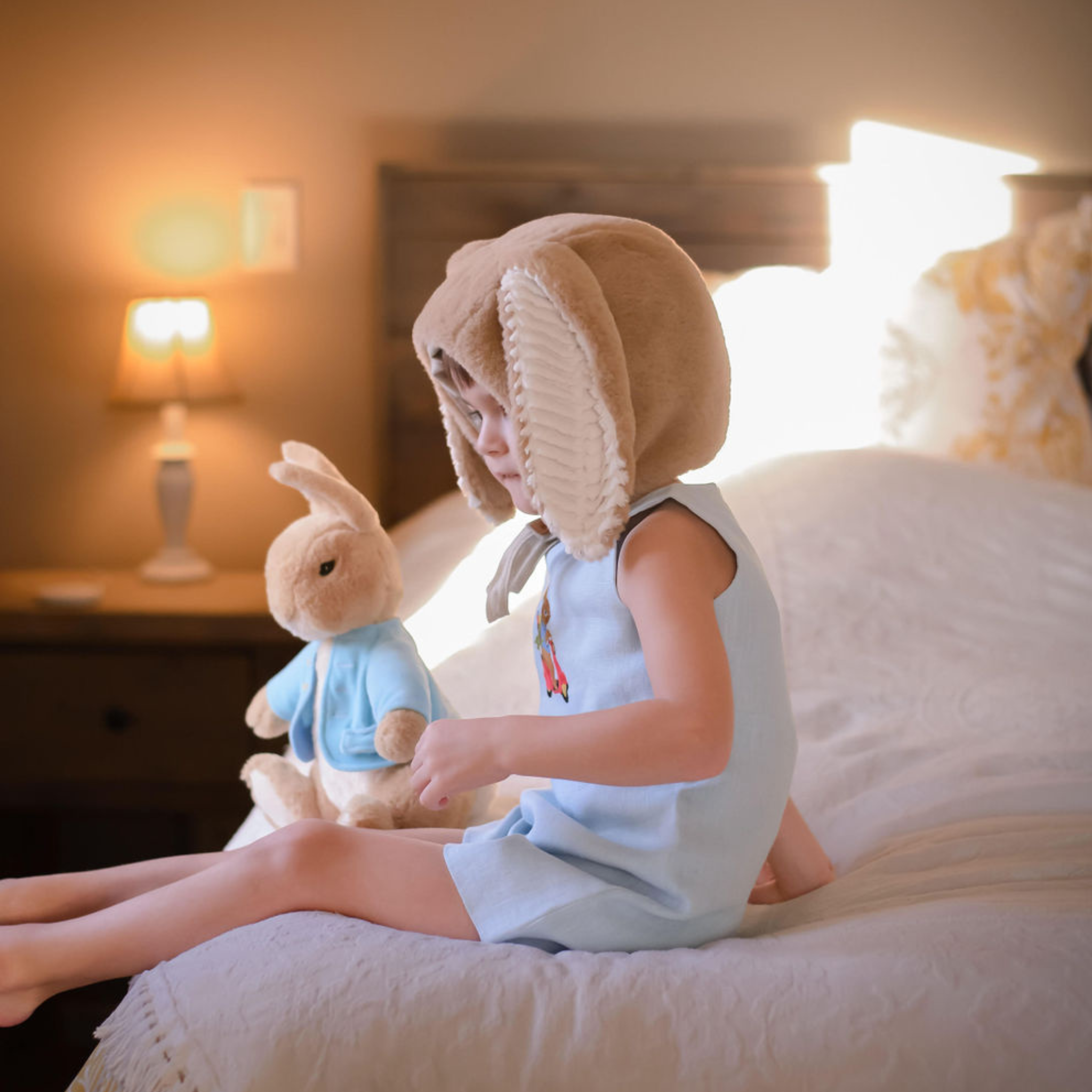 toddler wearing Scooter Bunny Linen Jon Jon sits on a bed with a plush bunny, showcasing the embroidered vintage-style outfit
