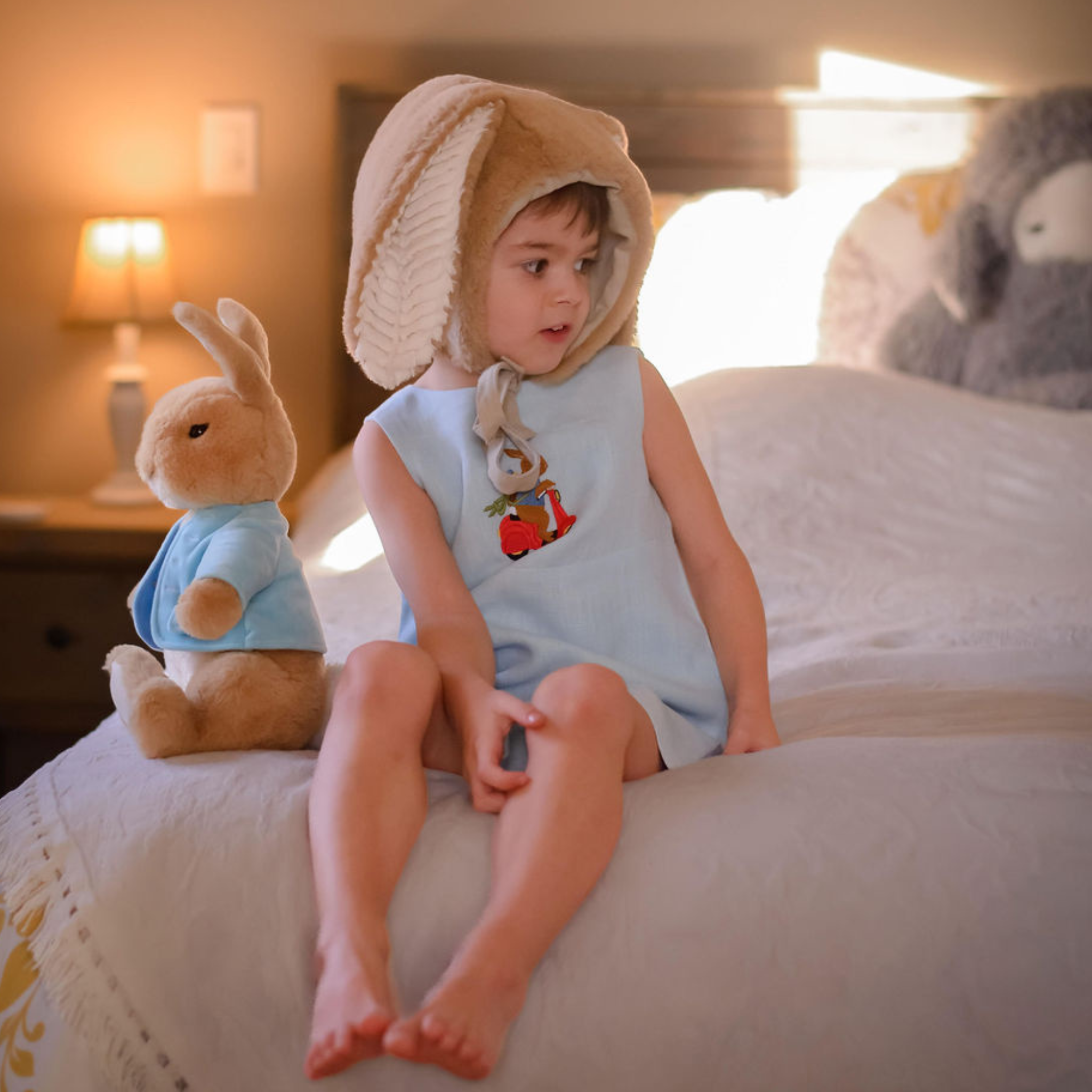 Toddler wearing light blue linen romper with embroidered scooter bunny, sitting on a bed with a plush bunny. Heirloom-quality baby clothing