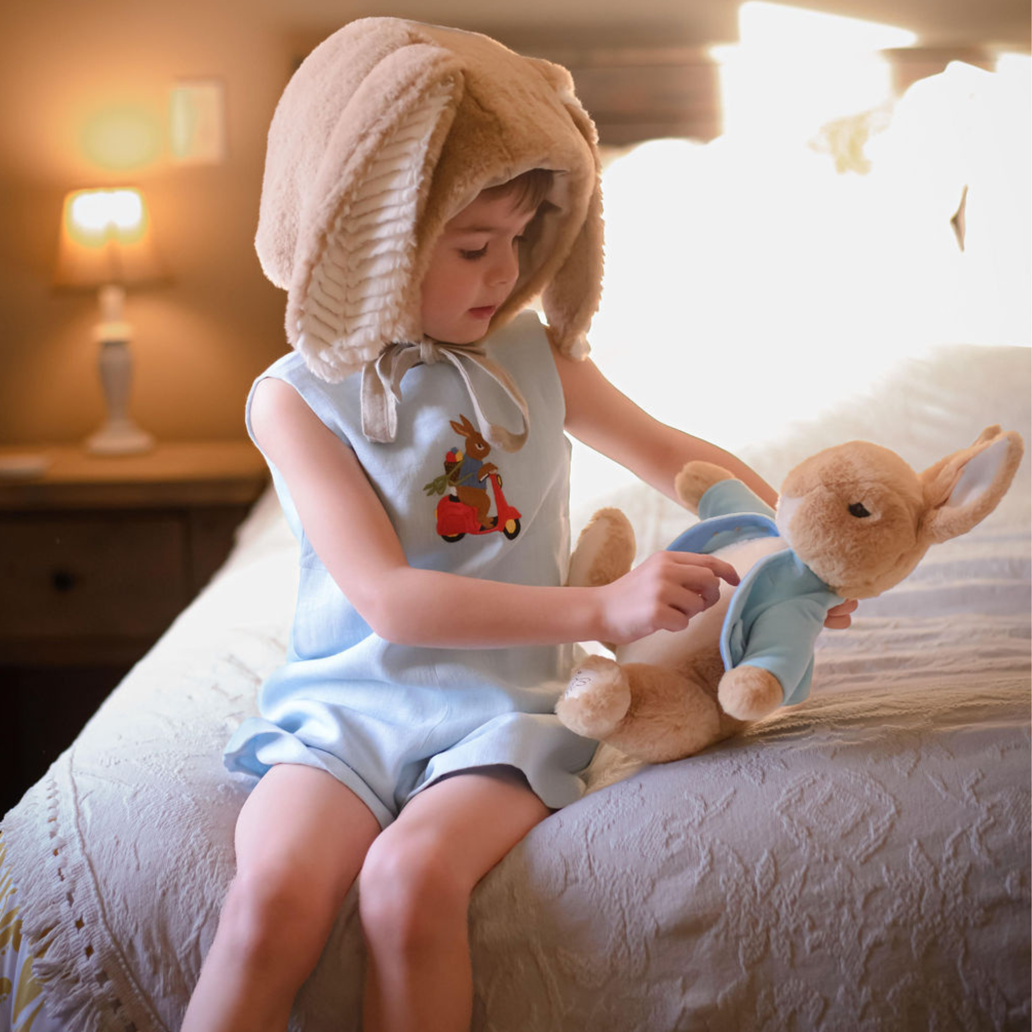 Toddler wearing a light blue linen romper with embroidered scooter bunny, sitting on a bed with a plush bunny. Heirloom-quality baby outfit