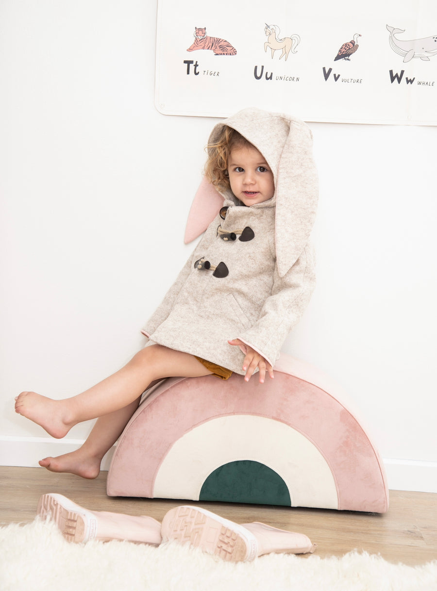 kid wearing a Snowshoe Rabbit Coat with bunny ears, toggle fasteners, and Merino wool fabric, sitting on a pastel rainbow cushion.