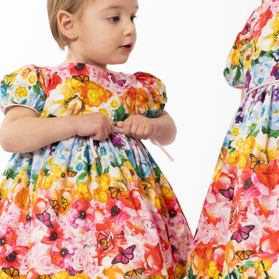 Toddler wearing Rainbow Bloom Dress with vibrant floral and butterfly print. Puff sleeves and a waist tie add charm to this colorful outfit
