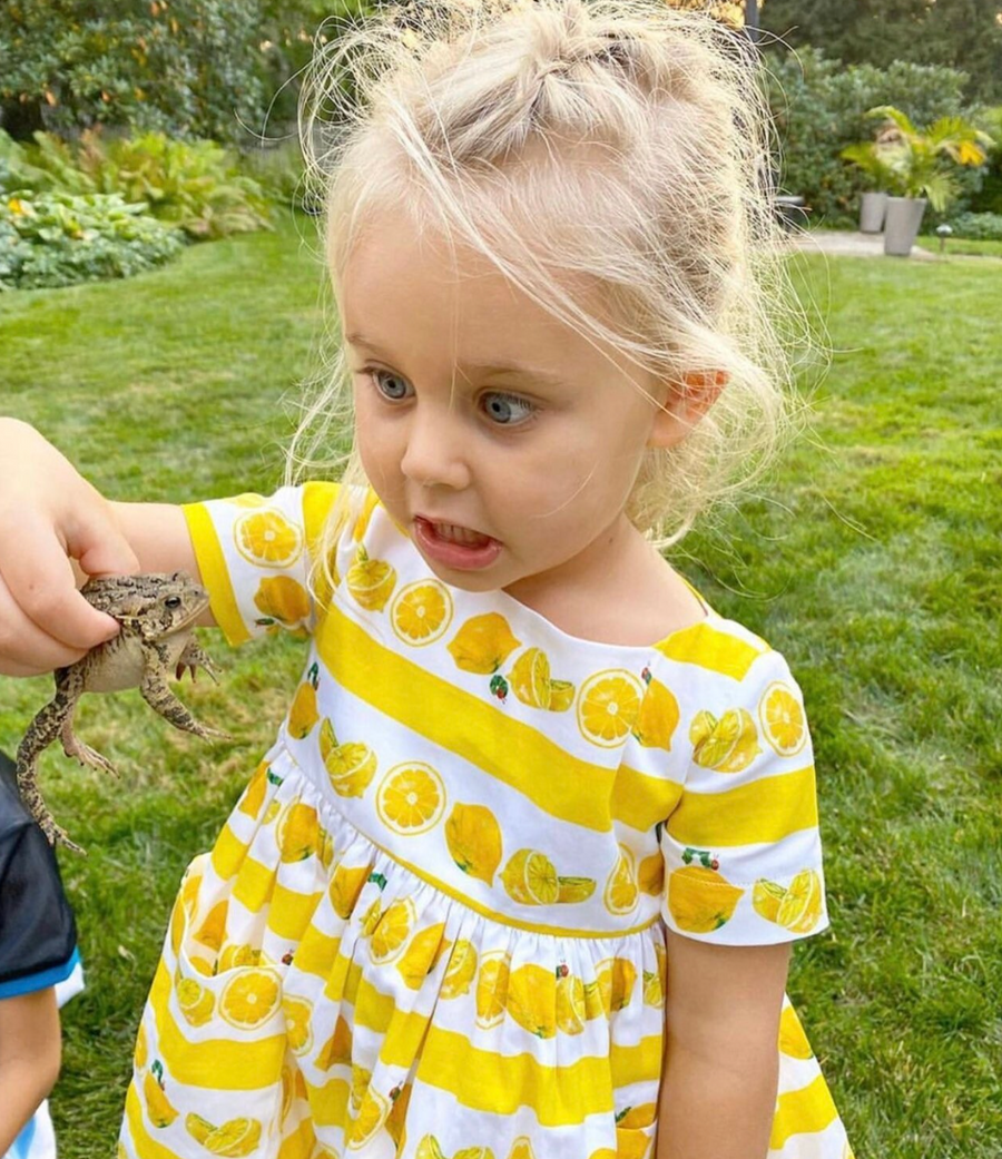 Young girl wearing a lemon-print dress, making a funny face while holding a lizard in a grassy backyard.