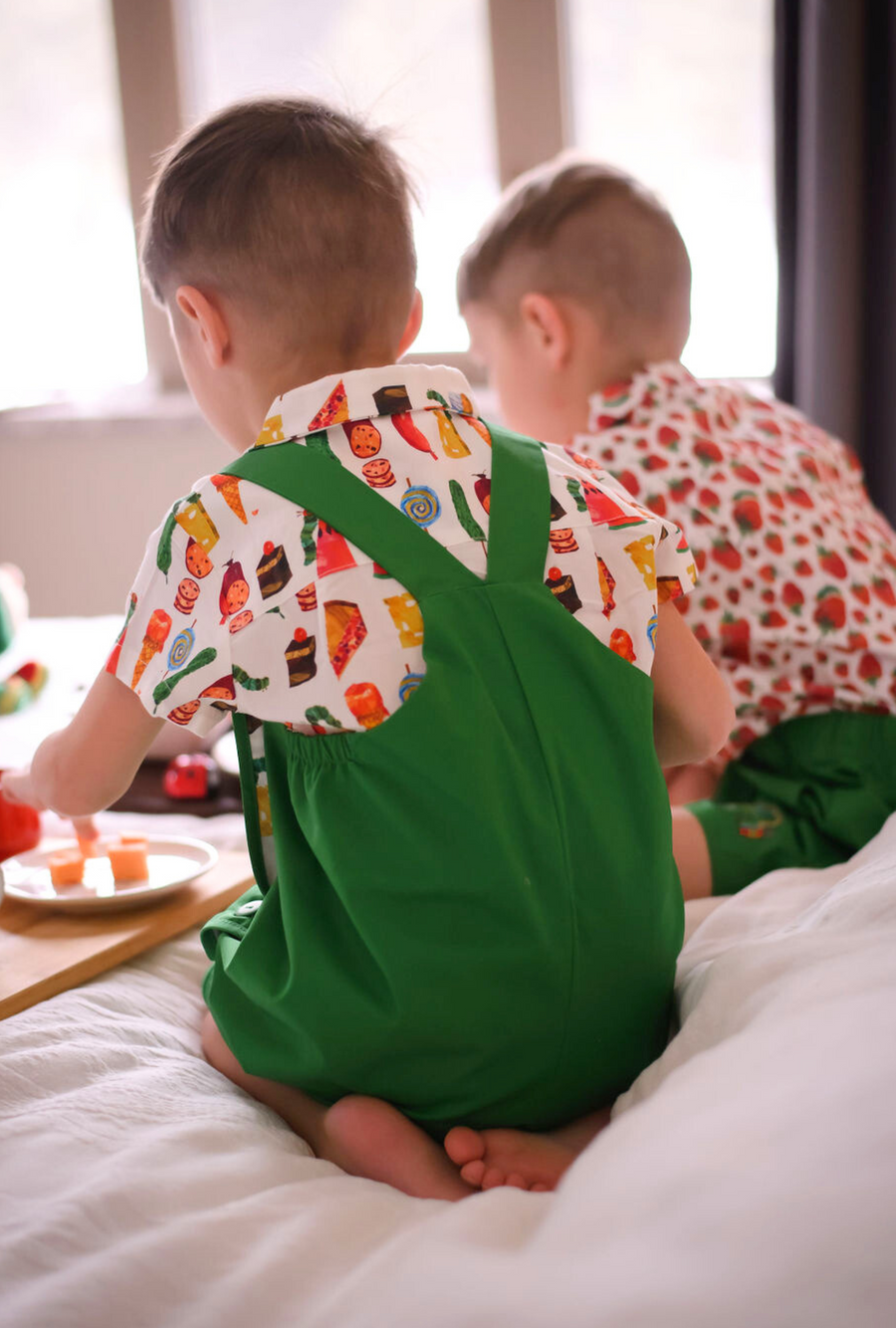 Back view of a toddler wearing a green Hungry Caterpillar romper with crossed straps and a food print shirt
