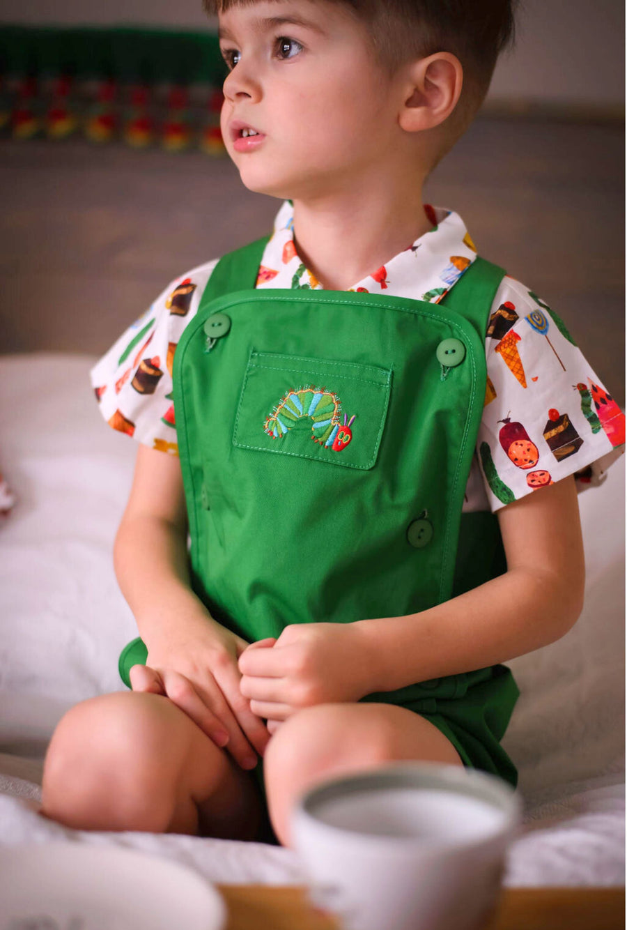 Toddler sitting on a bed wearing a green Hungry Caterpillar romper with embroidery, layered over a food print shirt