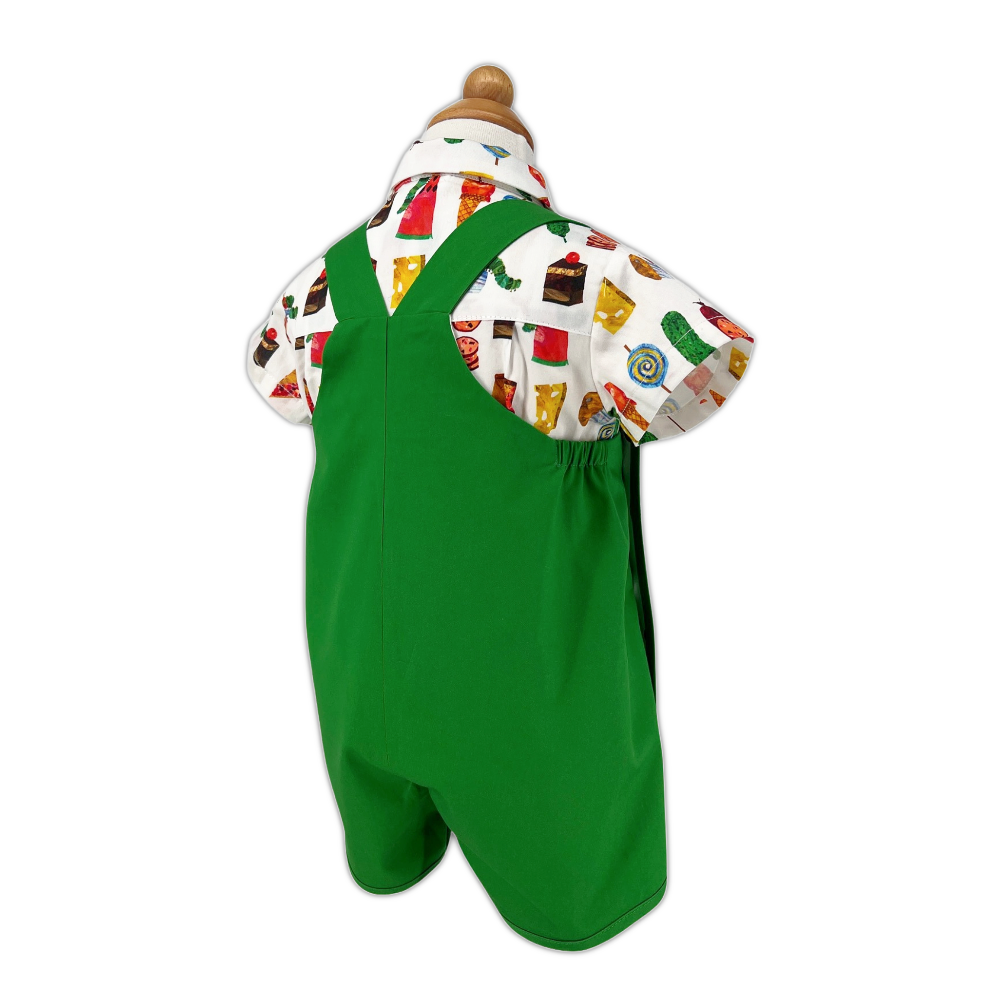 Back view of  Very Hungry Caterpillar romper displayed on form, showing green twill fabric, crisscross straps, and colorful food print shirt
