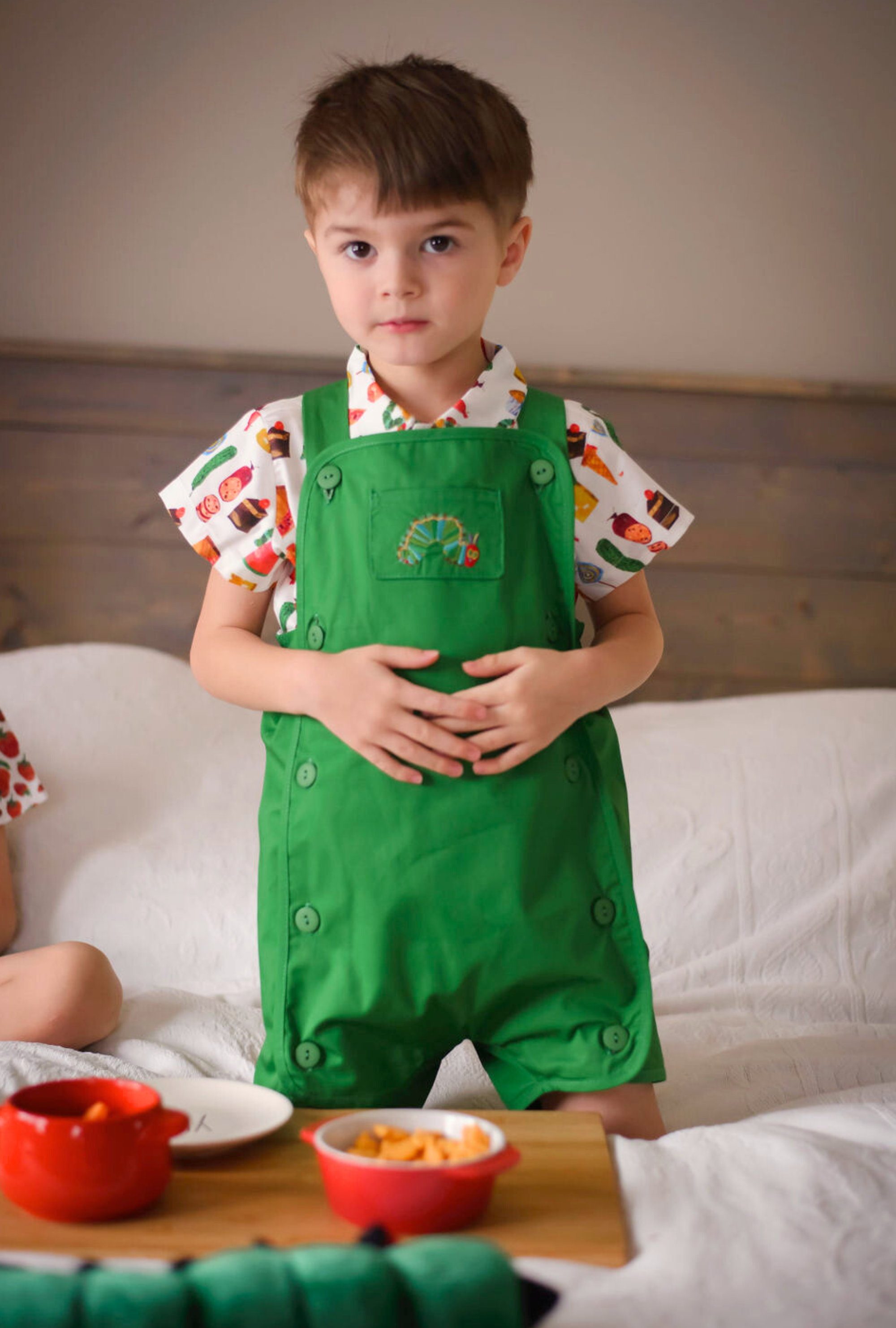 Toddler standing in a green Hungry Caterpillar romper with embroidered detail, layered over a food print shirt, with snack bowls on a wooden tray.