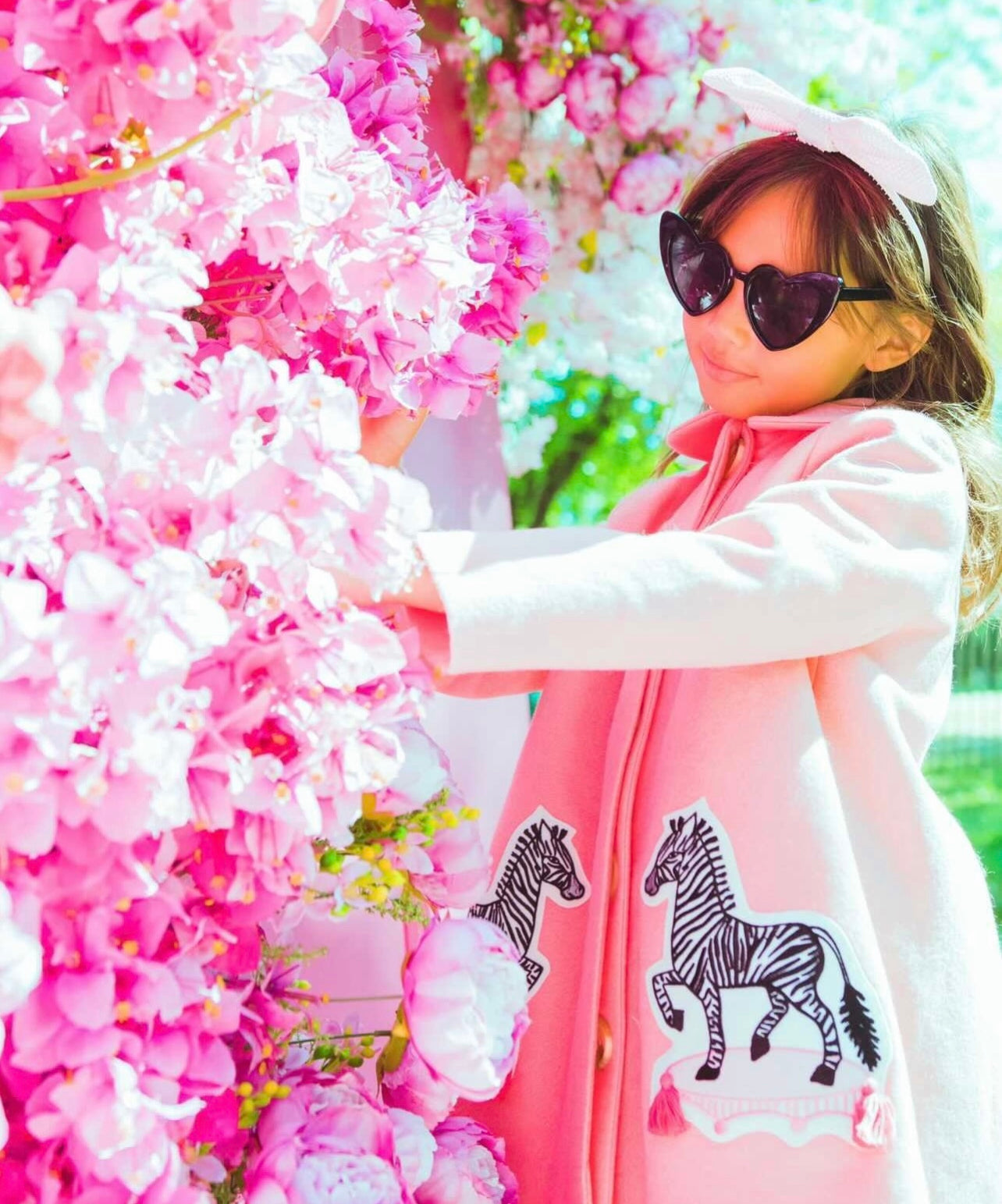 Young girl in a pink zebra-appliqué coat, accessorized with a bow and sunglasses, near blooming flower