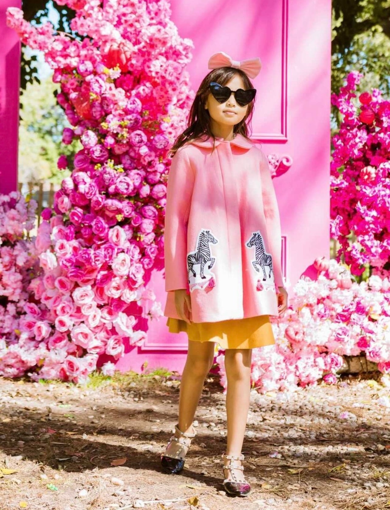 Girl wearing Yolanda and Tatty pink wool coat with zebra appliqués, styled with a skirt and bow, in a floral setting