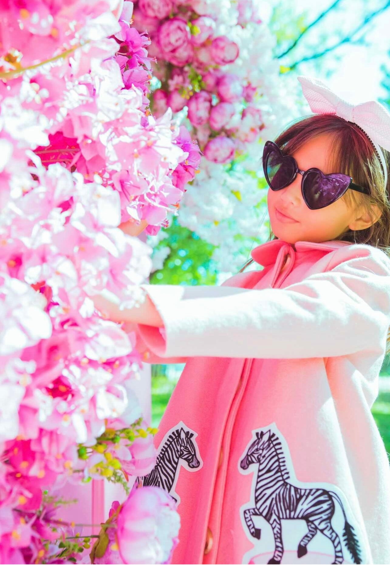 Girl wearing Yolanda and Tatty pink wool coat with zebra appliqués, styled with a skirt and bow, in a floral setting