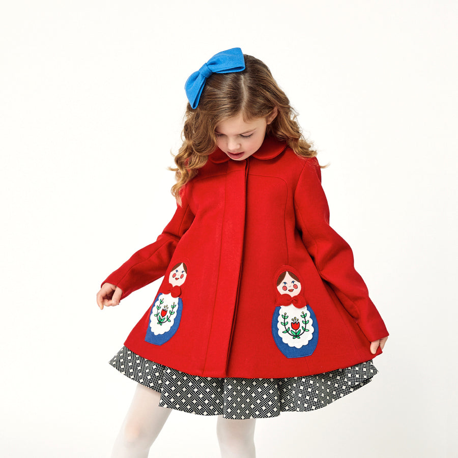 Close up of girl modeling red coat while wearing a polkadot dress underneath and a blue bow on top of her head.