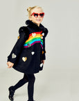 Girl modeling black coat with wearing black tights and shoes and her hair in two buns with red heart sunglasses on.