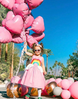 Little Little girl with heart sunglasses standing in front of palm trees holding big bunch of pink heart shaped balloons. She wears a pink sundress with a rainbow on the front, full skirt, and white peplum shaped like clouds.