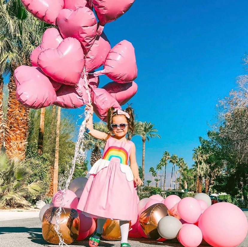 Little Little girl with heart sunglasses standing in front of palm trees holding big bunch of pink heart shaped balloons. She wears a pink sundress with a rainbow on the front, full skirt, and white peplum shaped like clouds.
