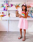 Little Older girl wearing a pink sundress with appliqued rainbow on the front, full skirt, and white peplum shaped like clouds.  She has a big pink bow in her hair and is holding pink cotton cangy, while standing in front of a white wall with shelves of cure stuffed toys. 