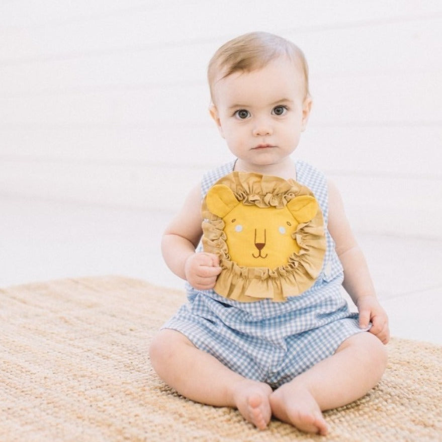 Baby wearing Blue Gingham Lion Romper for boys and girls with embroidered lion face and ruffled mane, sitting on a woven rug, looking at the camera