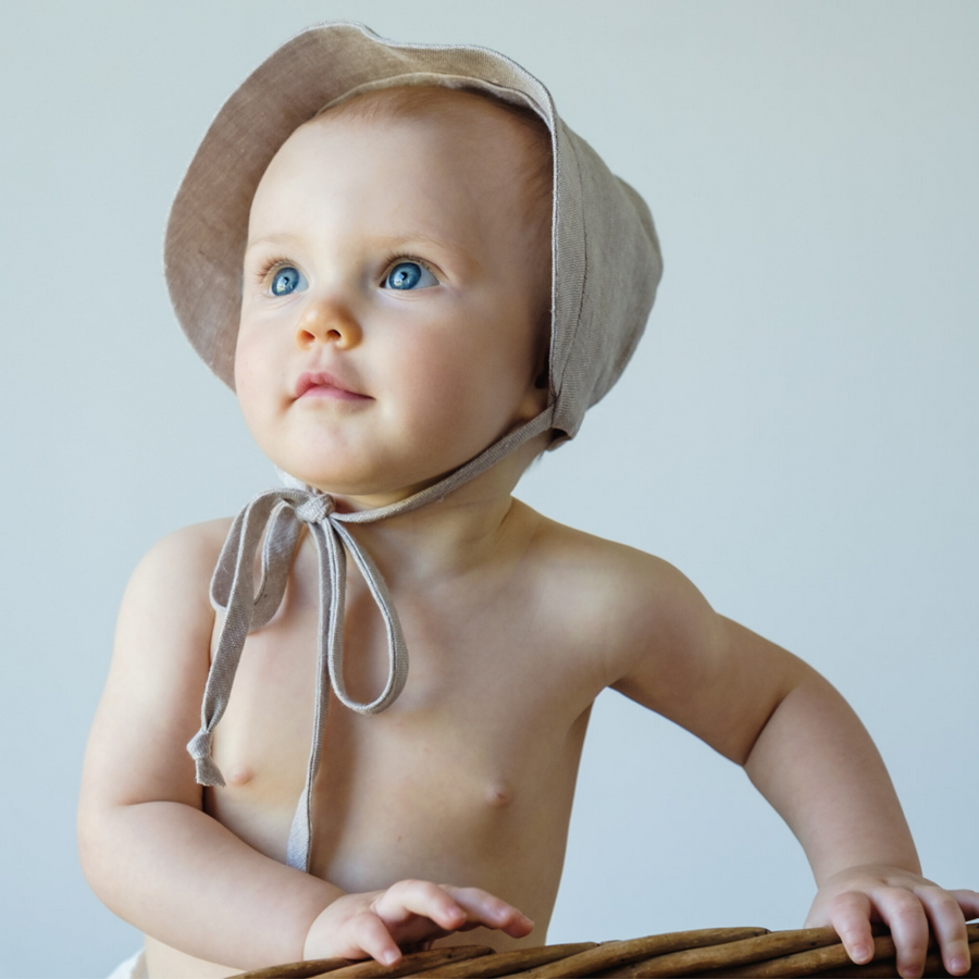 Baby wearing a flax linen wide-brim sun bonnet in wheat color with chin ties for sun protection.
