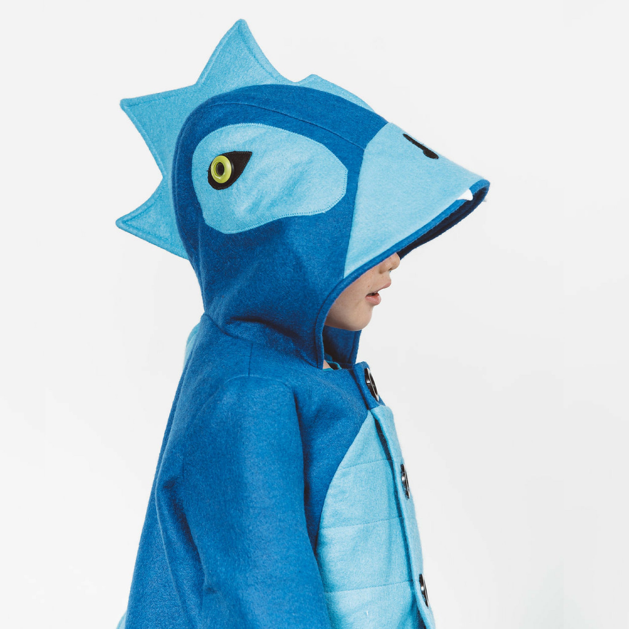 Side view of a young boy wearing the Blithe Blue Dinosaur Coat, featuring a playful dinosaur hood with spikes and a button-up front