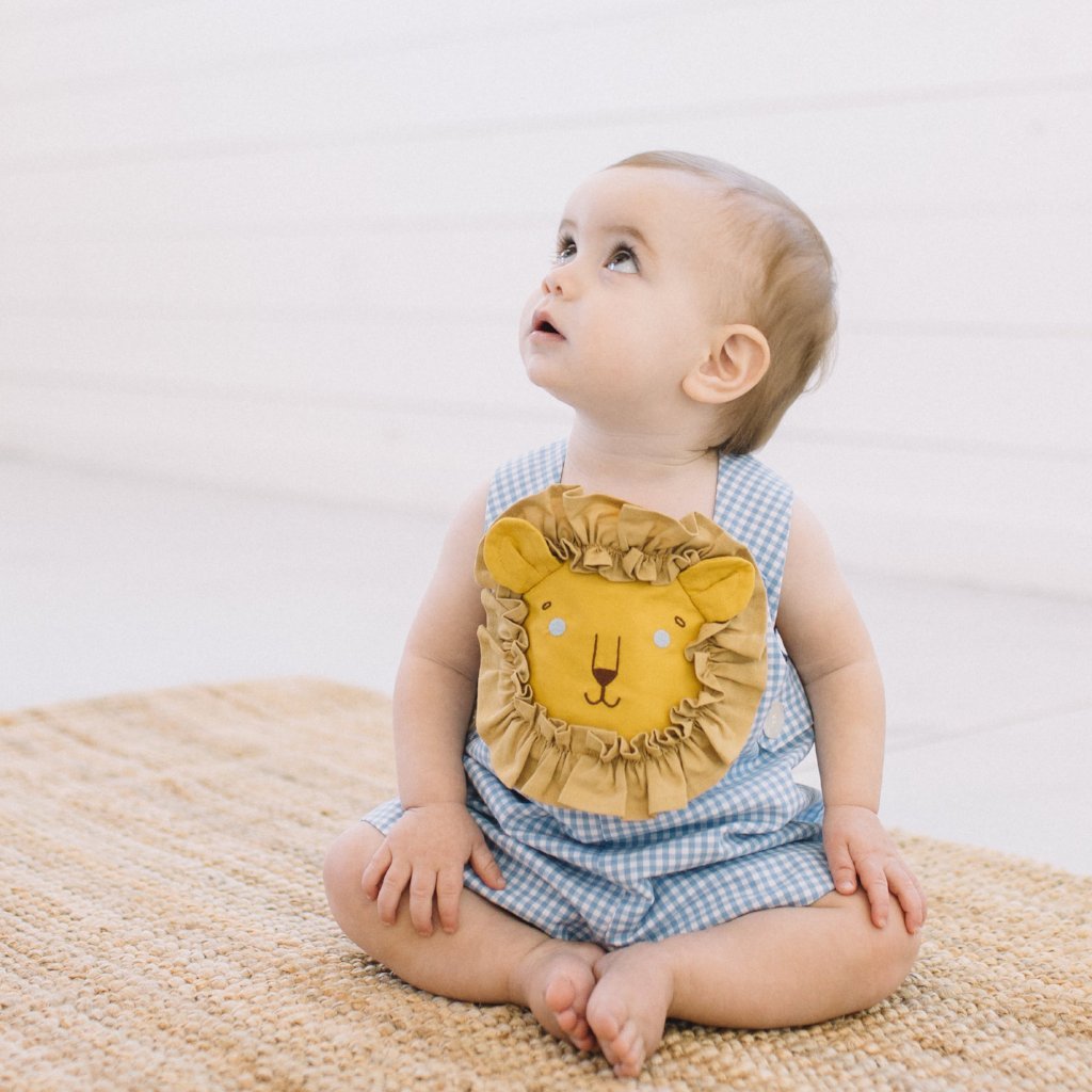 Baby in Blue Gingham Lion Romper sitting on a woven rug, gazing upward. Features lion face appliqué with ruffled mane. Childsware for boys and girls